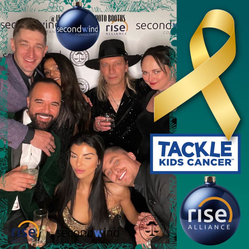 Group of adults posing closely together at a holiday photo booth, smiling and holding drinks, with Rise Alliance and Second Wind branding in the background. A gold ribbon and “Tackle Kids Cancer” logo appear on the right side of the image