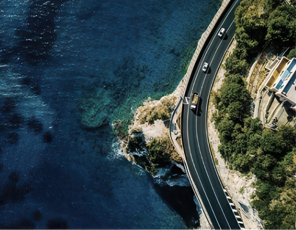 Aerial view of a winding coastal road hugging a rocky shoreline, with several cars driving beside clear blue ocean water and cliffs lined with greenery.